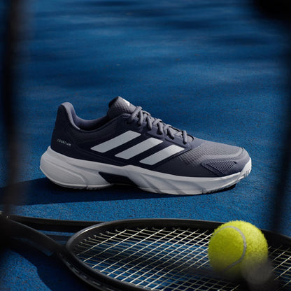 An adidas CourtJam Control 3 Men’s Tennis Shoe in Dark Blue and Cloud White rests on a blue court beside a black tennis racket and a yellow tennis ball, delivering enhanced stability as shadows stretch across the ground.