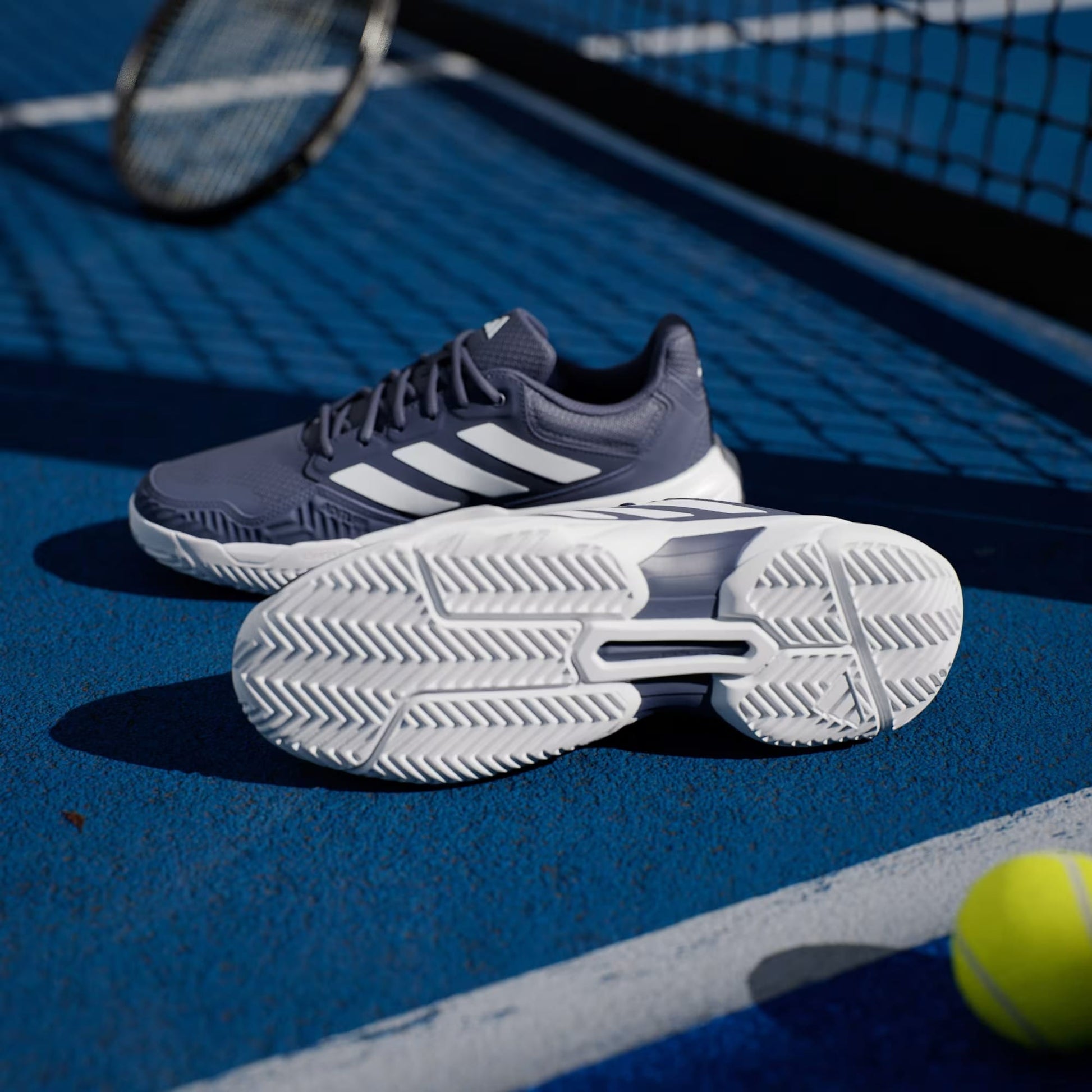 A pair of adidas CourtJam Control 3 Men’s Tennis Shoes in Dark Blue and Cloud White rest on a blue tennis court, with one shoe upright and the other displaying its sole to highlight their stability. In the background, a tennis ball and racket can be seen.