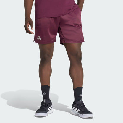 A person wearing adidas Climacool Ergo Pro Men’s Tennis Shorts in maroon, paired with a matching maroon shirt, black adidas socks, and black-and-white adidas sneakers stands with one hand in their pocket against a light background.