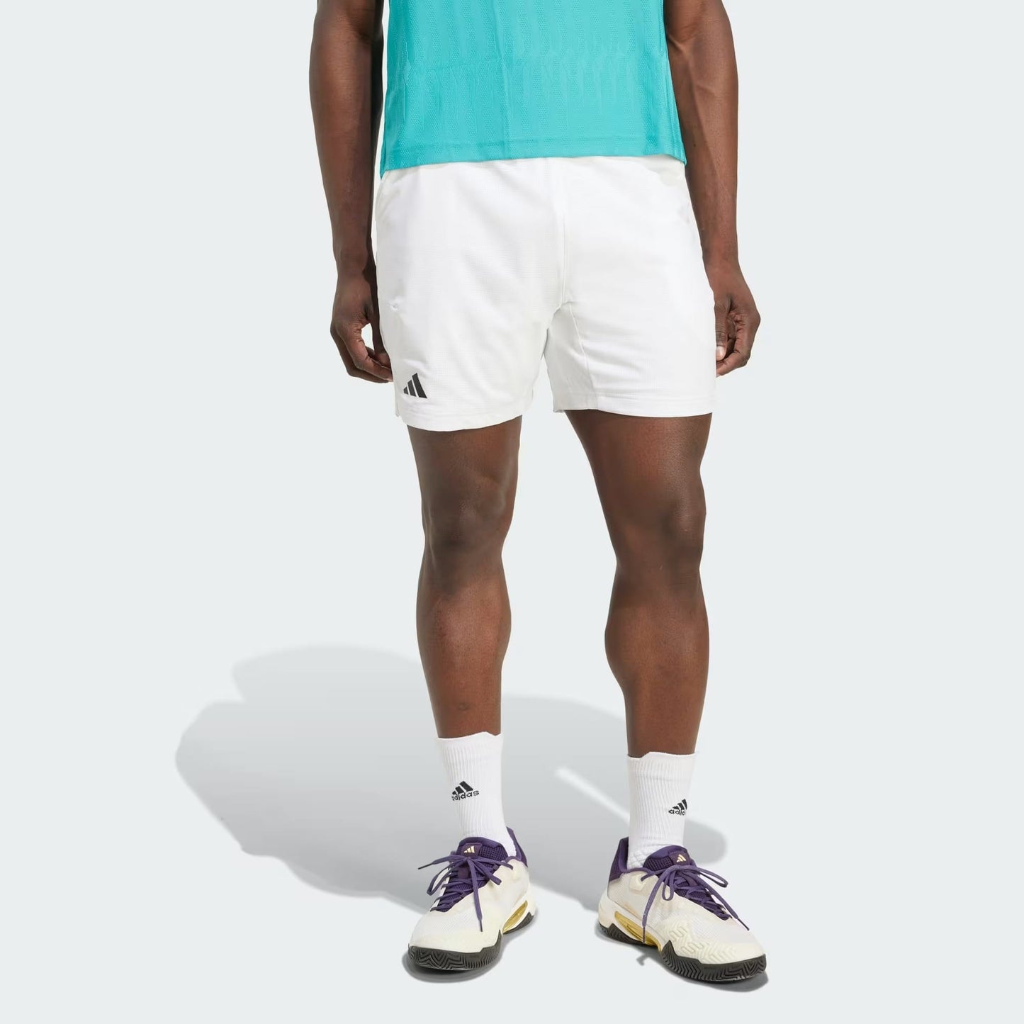 A person in a turquoise shirt, adidas Climacool Ergo Men's Tennis Shorts (White) by adidas, white crew socks, and white sneakers with purple and yellow accents stands against a plain light background.