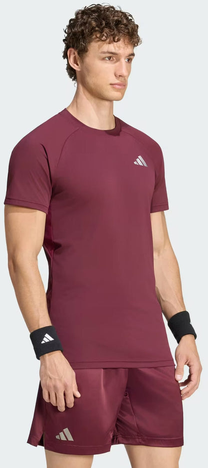 A man with curly hair is wearing the adidas Climacool+ Freelift Pro Men’s Tennis T-Shirt in maroon and matching shorts featuring white adidas logos, along with black adidas wristbands, standing against a plain light background.
