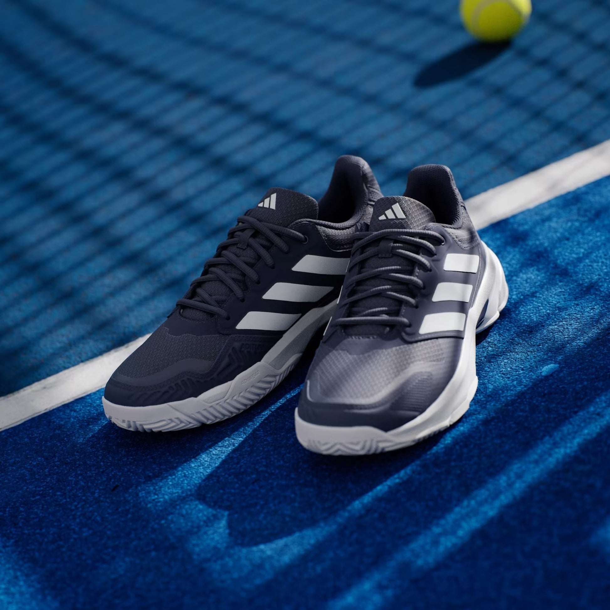 A pair of adidas CourtJam Control 3 Men’s Tennis Shoes in Dark Blue and Cloud White displayed on a blue tennis court, featuring enhanced stability, with a tennis ball and net seen in the background.