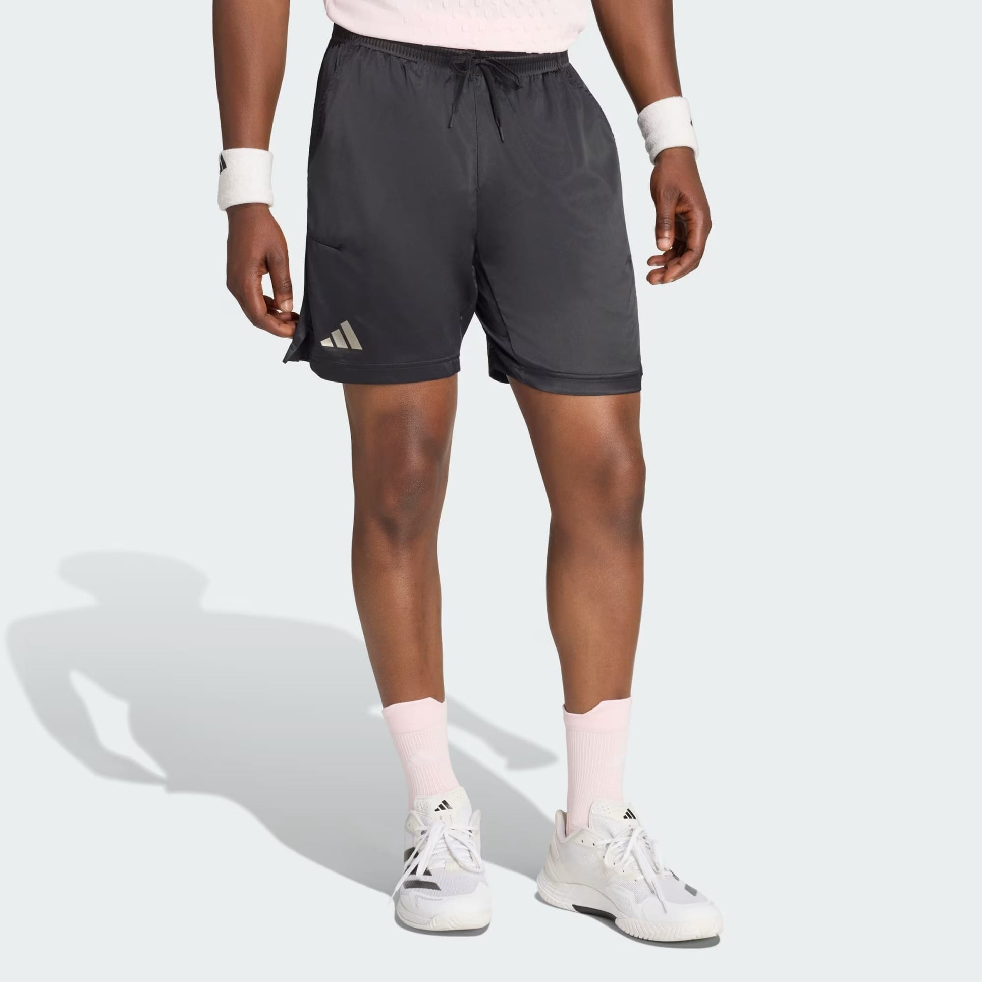 Wearing adidas Climacool Ergo Pro Men’s Tennis Shorts in black, along with a pink shirt, white Adidas sneakers, pink socks, and white wristbands, a person stands against a light background.