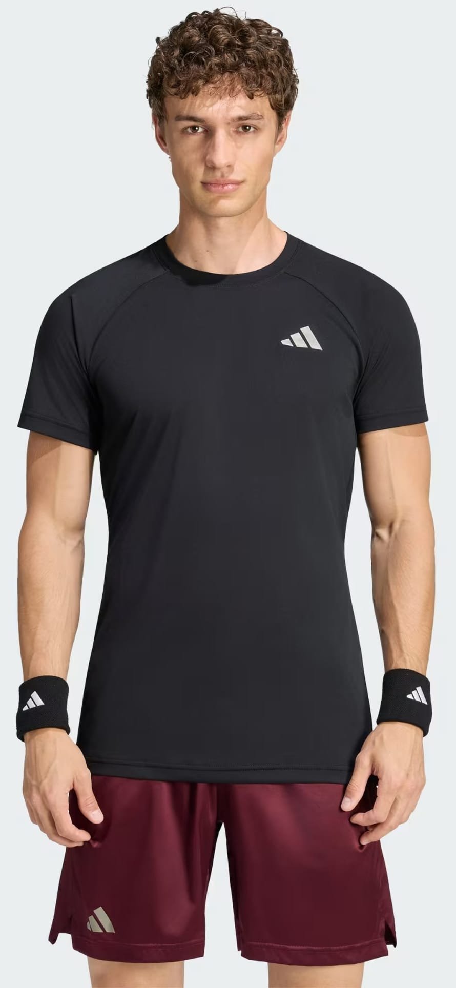 A man with curly hair wears an adidas Climacool+ Freelift Pro Men’s Tennis T-Shirt in black, maroon shorts, and black wristbands with white logos, standing facing forward against a plain light background.