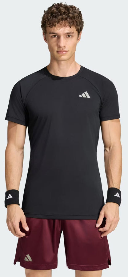A man with curly hair wears an adidas Climacool+ Freelift Pro Men’s Tennis T-Shirt in black, maroon shorts, and black wristbands with white logos, standing facing forward against a plain light background.