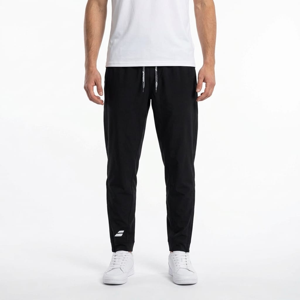 A person wearing a white t-shirt, Babolat Play Men’s Tennis Pants in black featuring a small white Babolat logo on the left leg, and white sneakers stands against a plain white background.