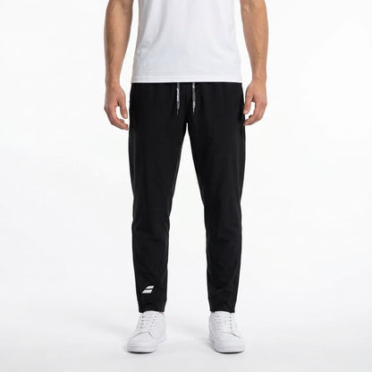 A person wearing a white t-shirt, Babolat Play Men’s Tennis Pants in black featuring a small white Babolat logo on the left leg, and white sneakers stands against a plain white background.