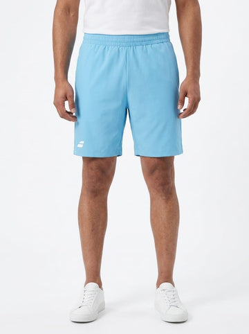 A person is wearing Babolat Play Mens Tennis Shorts in Cyan Blue, along with a white T-shirt and white sneakers, standing against a plain white background. Only the lower torso and legs are visible.