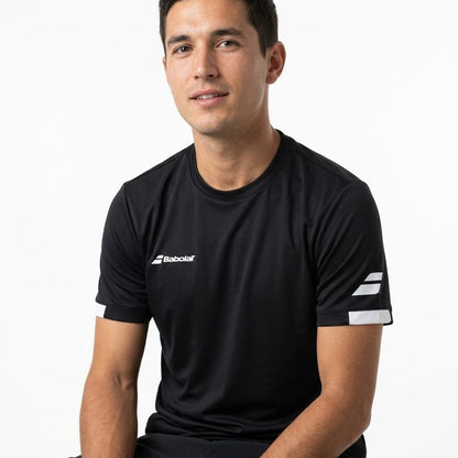 A man with short dark hair sits against a plain white background, wearing the Babolat Play Men’s Crew Neck Tennis T-Shirt in black. This classic crew neck from Babolat features white sleeve accents and is crafted from recycled polyester, blending style and sustainability as he looks at the camera with a slight smile.