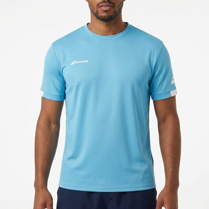 A man stands against a plain white background wearing the Babolat Play Men’s Crew Neck Tennis T-Shirt in cyan blue, which features short sleeves and is made from recycled polyester, paired with dark shorts.