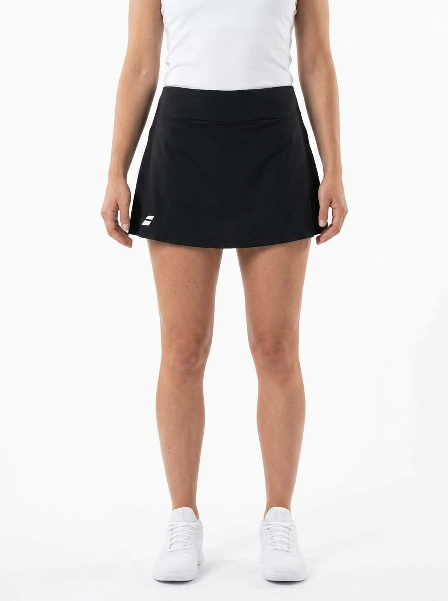 A person stands against a white background, wearing a white sleeveless top and the Babolat Play Women’s Tennis Skirt in black by Babolat, featuring integrated shorts and a small white logo. The skirt is crafted from recycled polyester. They have paired it with white sneakers, with their arms relaxed at their sides.