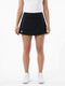 A person stands against a white background, wearing a white sleeveless top and the Babolat Play Women’s Tennis Skirt in black by Babolat, featuring integrated shorts and a small white logo. The skirt is crafted from recycled polyester. They have paired it with white sneakers, with their arms relaxed at their sides.