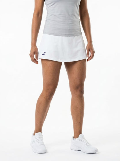 A person is wearing a gray sleeveless top, the Babolat Play Womens Tennis Skirt in white made from recycled polyester and featuring a small dark Babolat logo, along with white athletic shoes, while standing against a plain white background.