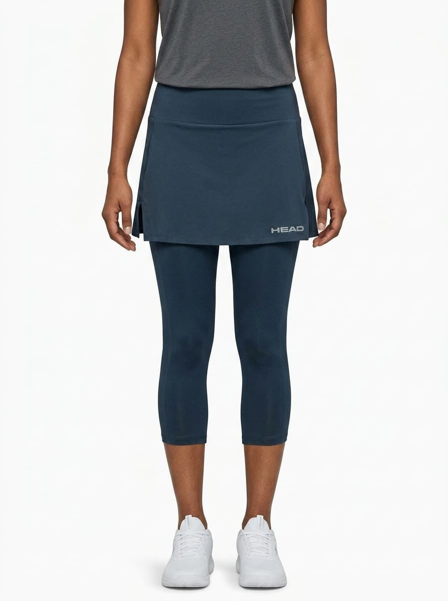 A woman stands against a plain white background wearing a grey short-sleeve top, white sneakers, and the HEAD Club 3/4 Tights Women’s Tennis Skort in navy. The skort, featuring the HEAD logo, is crafted from moisture transfer microfibre to keep you cool and comfortable on and off the court.