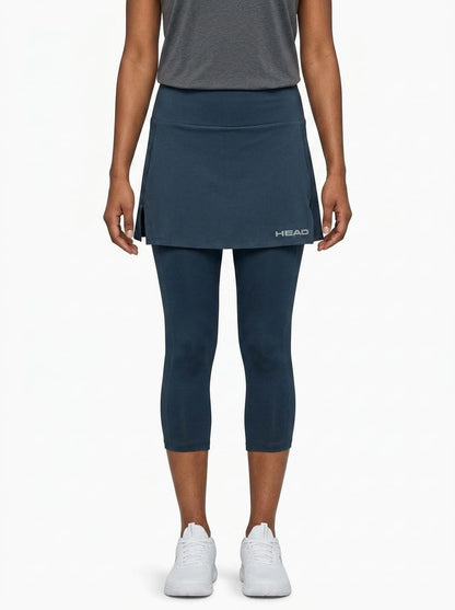 A woman stands against a plain white background wearing a grey short-sleeve top, white sneakers, and the HEAD Club 3/4 Tights Women’s Tennis Skort in navy. The skort, featuring the HEAD logo, is crafted from moisture transfer microfibre to keep you cool and comfortable on and off the court.