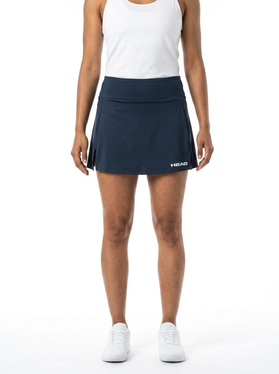 A woman wearing a white tank top and the HEAD Club Basic Women’s Tennis Skort Long in navy, featuring moisture-wicking fabric and the signature “HEAD” logo, paired with white sneakers. She stands against a plain white background with her arms relaxed at her sides.