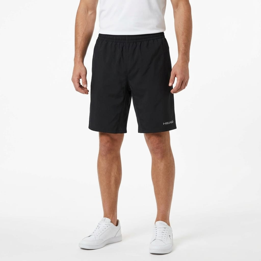 A person wearing a white shirt, HEAD Club Bermudas Men’s Tennis Shorts in black, and white sneakers stands against a plain white background.