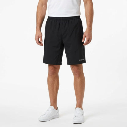 A person wearing a white shirt, HEAD Club Bermudas Men’s Tennis Shorts in black, and white sneakers stands against a plain white background.