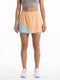 A woman stands against a white background, wearing a taupe crop top and the HEAD Dynamic Womens Tennis Skort in Apricot and Light Blue. She completes her look with white sneakers. The skort by HEAD is crafted with moisture transfer microfibre and 4-way stretch for exceptional comfort and style.
