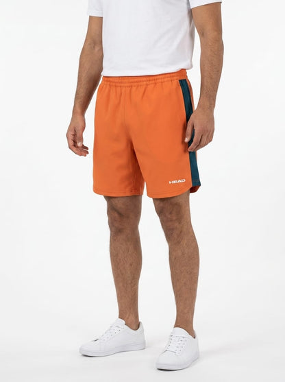 A person wearing a white t-shirt, HEAD Power Mens Tennis Shorts - CN by HEAD in orange with a teal stripe featuring 4-way stretch and moisture wicking fabric, and white sneakers stands against a plain white background. The image is cropped just below the shoulders.