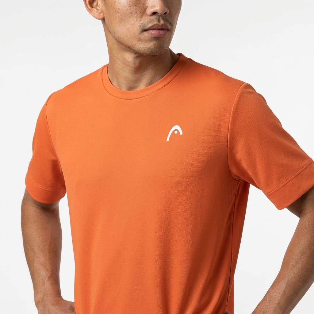 A man stands with his hands on his hips against a plain white background, wearing the HEAD Slice II Mens Tennis T-Shirt in orange. This short-sleeve t-shirt by HEAD features a small white logo on the chest and is designed with moisture-wicking fabric to keep you comfortable both on and off the court.