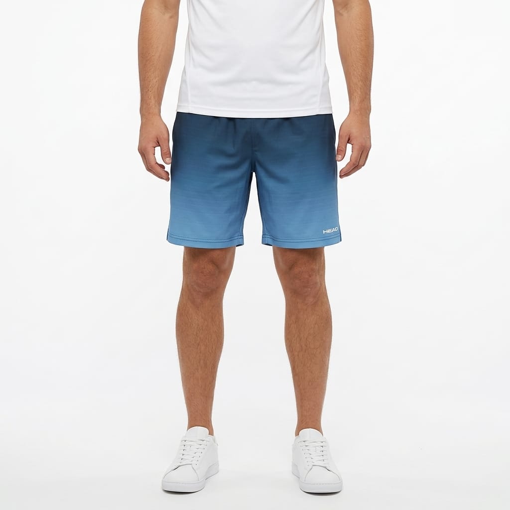 A man stands against a plain white background with his arms relaxed at his sides, wearing a white t-shirt, white sneakers, and HEAD Vision Power II Mens Tennis Shorts in Navy/Blue by HEAD, which feature 4-way stretch and moisture transfer microfiber.