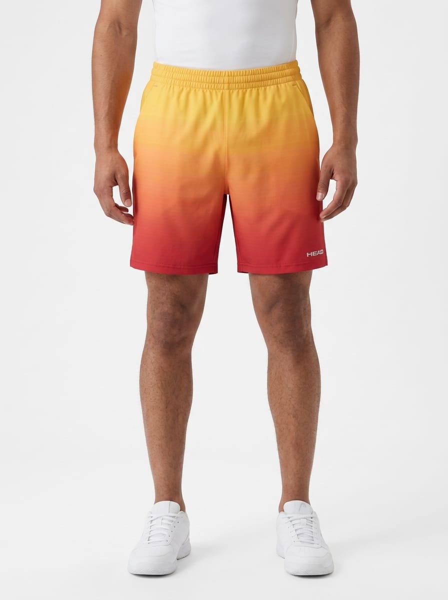 A person wearing white sneakers and HEAD Vision Power II Mens Tennis Shorts by HEAD, featuring a striking gradient that fades from yellow at the top to red at the bottom. The background is plain white.