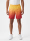 A person wearing white sneakers and HEAD Vision Power II Mens Tennis Shorts by HEAD, featuring a striking gradient that fades from yellow at the top to red at the bottom. The background is plain white.