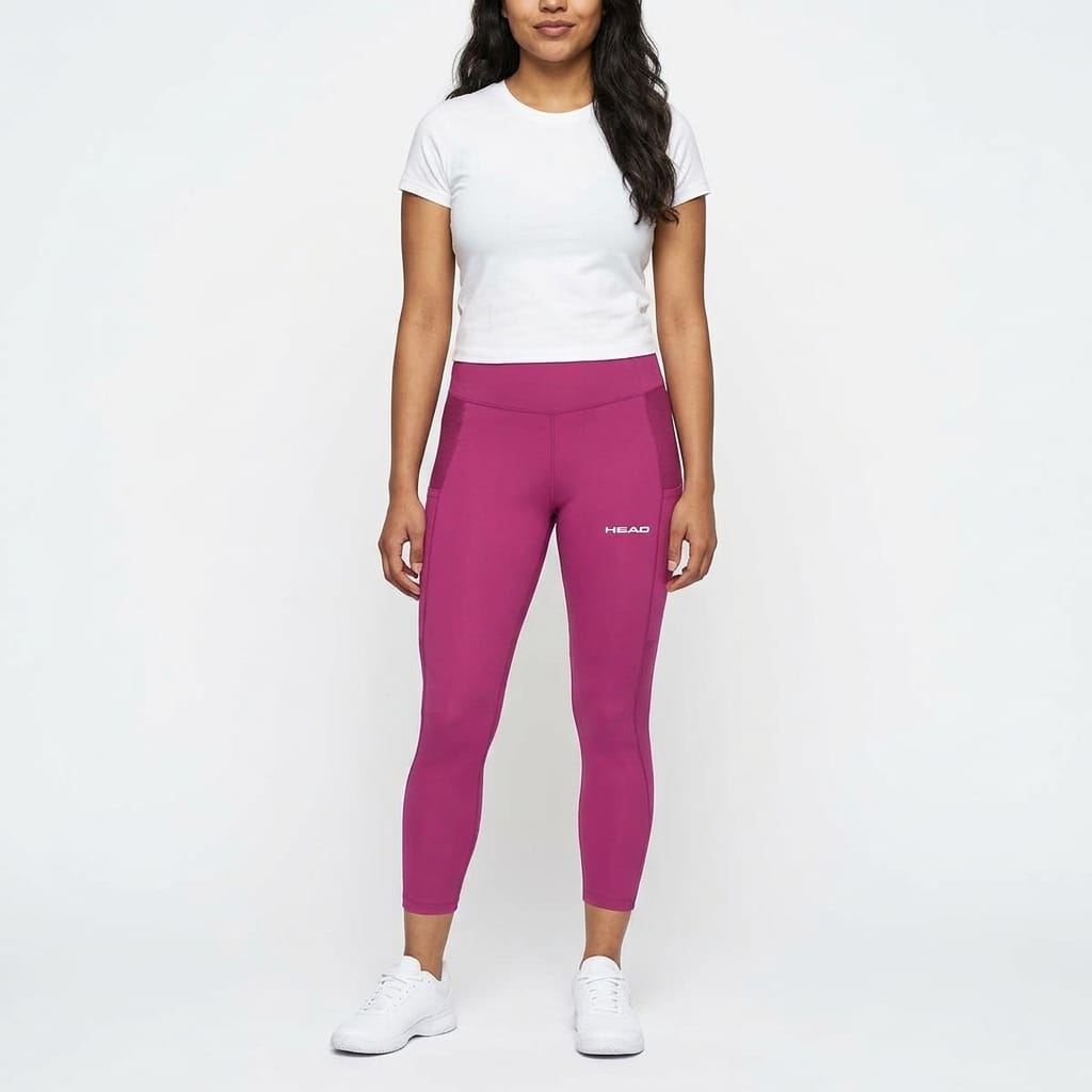 A woman stands against a white background wearing a white T-shirt, vivid pink HEAD Vision Women’s Tech Tennis Tights Leggings with the HEAD logo, and white sneakers. She has long dark hair and her arms rest by her sides.