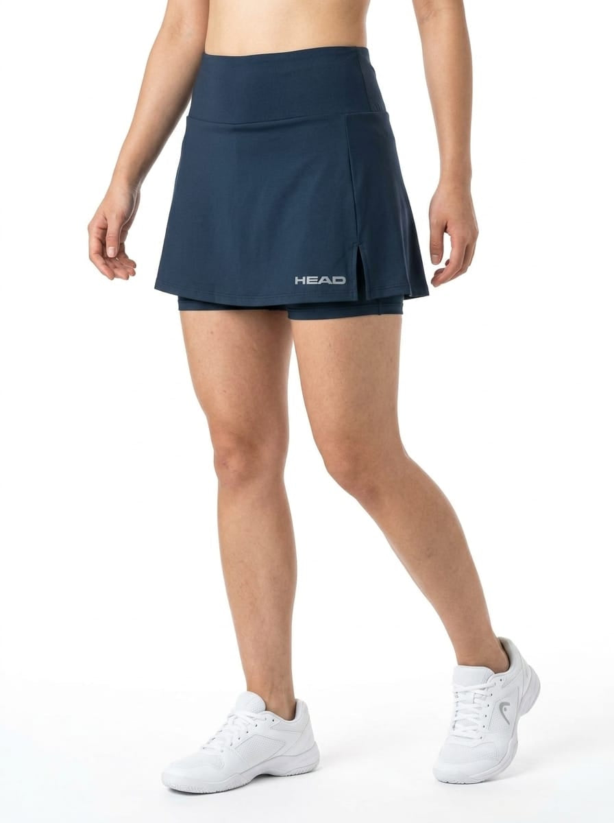 A person wearing the HEAD Womens Club Basic Tennis Skort - Navy by HEAD—a quick-dry tennis skort featuring moisture transfer microfibre—paired with white sneakers and a bare midriff, stands against a plain white background.
