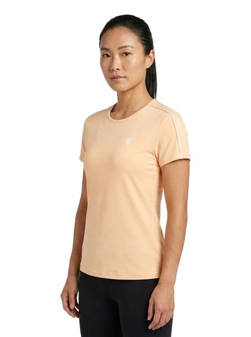 A woman with long dark hair in a ponytail wears the K-Swiss Hypercourt Crew Tee 3 Women’s Tennis T-Shirt in Peach Fuzz, paired with black pants, standing against a plain white background.