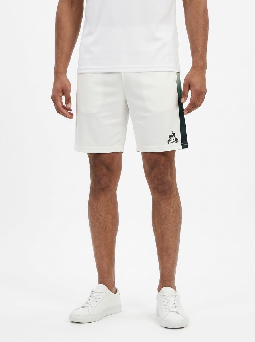 A man wearing a white t-shirt, Le Coq Sportif Pro Mens Tennis Shorts in Marshmallow—with a dark side stripe—and white sneakers stands against a plain white background. Designed by Le Coq Sportif for optimal comfort and movement, these breathable shorts are the ideal men’s tennis shorts for any active occasion.