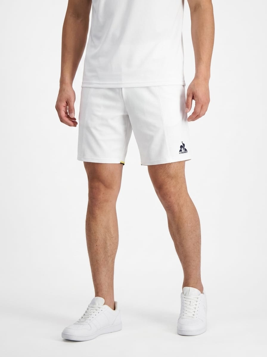 A person wearing a white t-shirt, Le Coq Sportif Pro Men’s Tennis Shorts in Optical White featuring a small blue logo on the left leg, and white sneakers stands against a plain white background.
