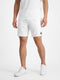 A person wearing a white t-shirt, Le Coq Sportif Pro Men’s Tennis Shorts in Optical White featuring a small blue logo on the left leg, and white sneakers stands against a plain white background.