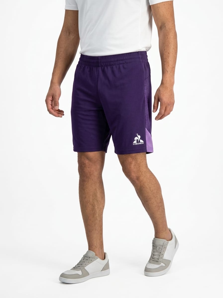 A person wearing a white T-shirt, Le Coq Sportif Pro Men’s Tennis Shorts in Purple Velvet, and gray and white sneakers stands against a plain white background.
