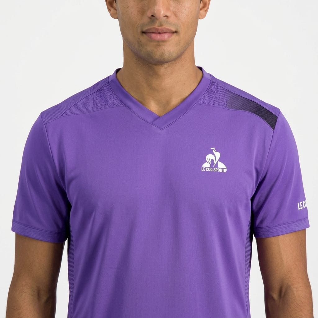 A person is wearing the Le Coq Sportif Pro Men’s Tennis T-Shirt in Chive Blossom, featuring short sleeves, a V-neck, and the iconic logo on the left chest, while standing against a plain light background. This breathable Le Coq Sportif sports shirt effortlessly blends comfort and style for any active occasion.