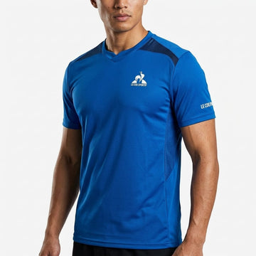 A man is wearing the Le Coq Sportif Pro Mens Tennis T-Shirt in Lapis Blue, featuring the “Le Coq Sportif” logo on the chest and left sleeve, while standing against a plain white background. This performance t-shirt from Le Coq Sportif offers a breathable fit, making it ideal for active play.