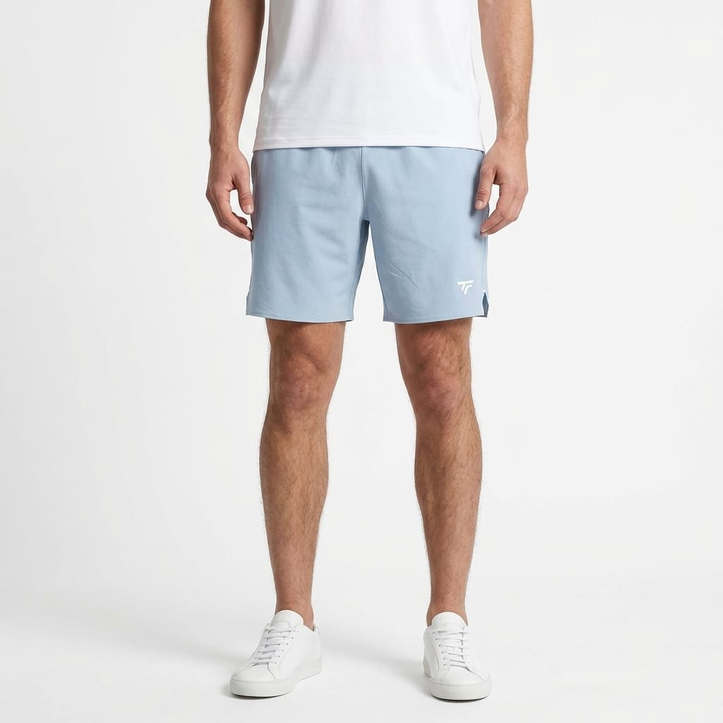 A person is wearing a white t-shirt, white sneakers, and Tecni­fibre Team Stretch Men’s Tennis Shorts in Glacier Blue, which feature a small white logo and stretch fabric for comfort, standing against a plain white background.