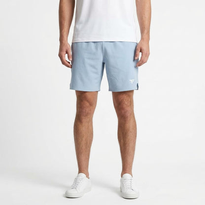 A person is wearing a white t-shirt, white sneakers, and Tecni­fibre Team Stretch Men’s Tennis Shorts in Glacier Blue, which feature a small white logo and stretch fabric for comfort, standing against a plain white background.