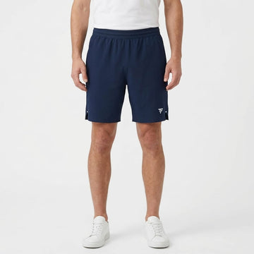A person wearing a white t-shirt, Tecnifibre Team Stretch Men’s Tennis Shorts in Marine Blue, and white sneakers stands against a plain white background.