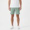 A person wearing a white t-shirt, Tecnifibre Team Stretch Men’s Tennis Shorts in sage green, and white sneakers stands against a plain white background.