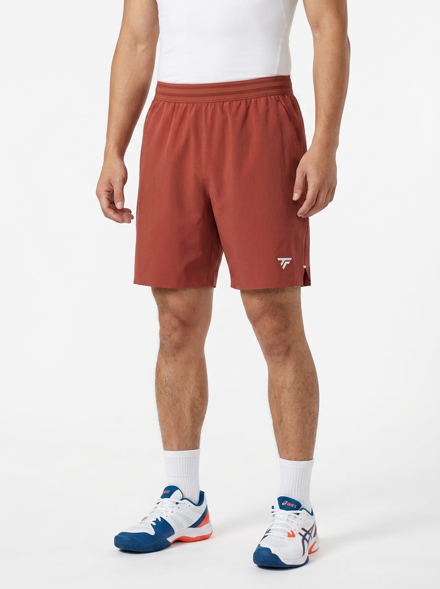 A man wearing a white tank top, Tecnifibre Team Stretch Mens Tennis Shorts in Terracotta, white socks, and white sneakers with blue and red accents stands against a plain white background.