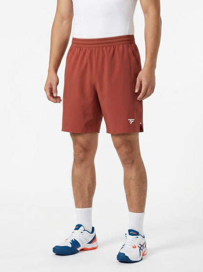 A man wearing a white tank top, Tecnifibre Team Stretch Mens Tennis Shorts in Terracotta, white socks, and white sneakers with blue and red accents stands against a plain white background.