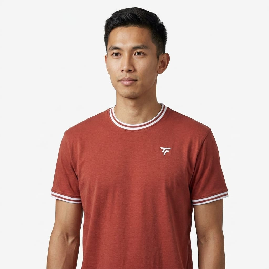 A man stands against a plain white background, looking slightly to his left, wearing the Tecnifibre Team Stretch Men’s Tennis T-Shirt in terracotta by Tecnifibre, which features white trim and a small logo.
