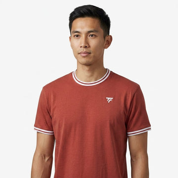 A man stands against a plain white background, looking slightly to his left, wearing the Tecnifibre Team Stretch Men’s Tennis T-Shirt in terracotta by Tecnifibre, which features white trim and a small logo.