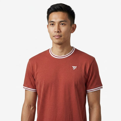 A man stands against a plain white background, looking slightly to his left, wearing the Tecnifibre Team Stretch Men’s Tennis T-Shirt in terracotta by Tecnifibre, which features white trim and a small logo.