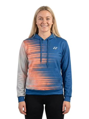 A person with long blonde hair smiles while wearing the Yonex YHD2025 Unisex Tennis Hoodie - Sunset, featuring a blue and orange gradient design in advanced Poly-Tech Knit fabric with a white Yonex logo on the chest, paired with black pants, standing against a plain white background.