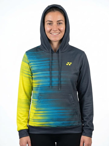 A woman smiles while wearing the Yonex YHD2025 Unisex Tennis Hoodie in Teal/Lime, featuring blue, yellow, and black gradient stripes with a small yellow Yonex logo on the chest, standing against a plain white background. This quick-dry hoodie is crafted from advanced Poly-Tech Knit fabric for superior comfort and performance.