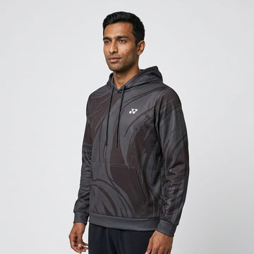 A man stands facing slightly left, wearing the Yonex YHD6 Unisex Tennis Hoodie - Storm in a black and gray pattern with a small white Yonex logo on the chest, against a plain light gray background.