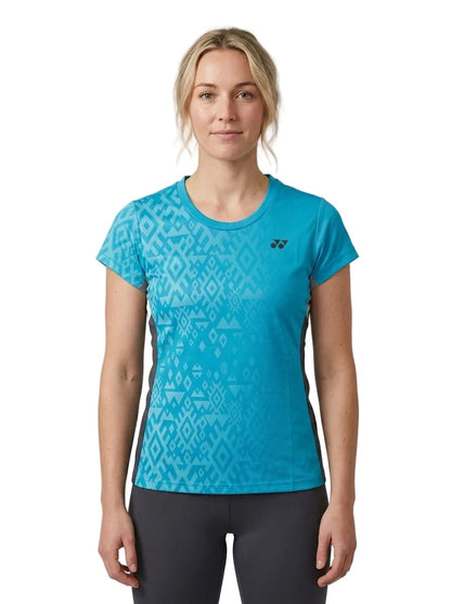 A woman stands facing forward with a neutral expression against a white background, wearing the Yonex YTL11 Women’s Tennis T-Shirt in Blue Sea—a short-sleeve, blue patterned top paired with dark pants. Made from lightweight performance fabric, this quick-drying Yonex shirt offers comfort both on and off the court.